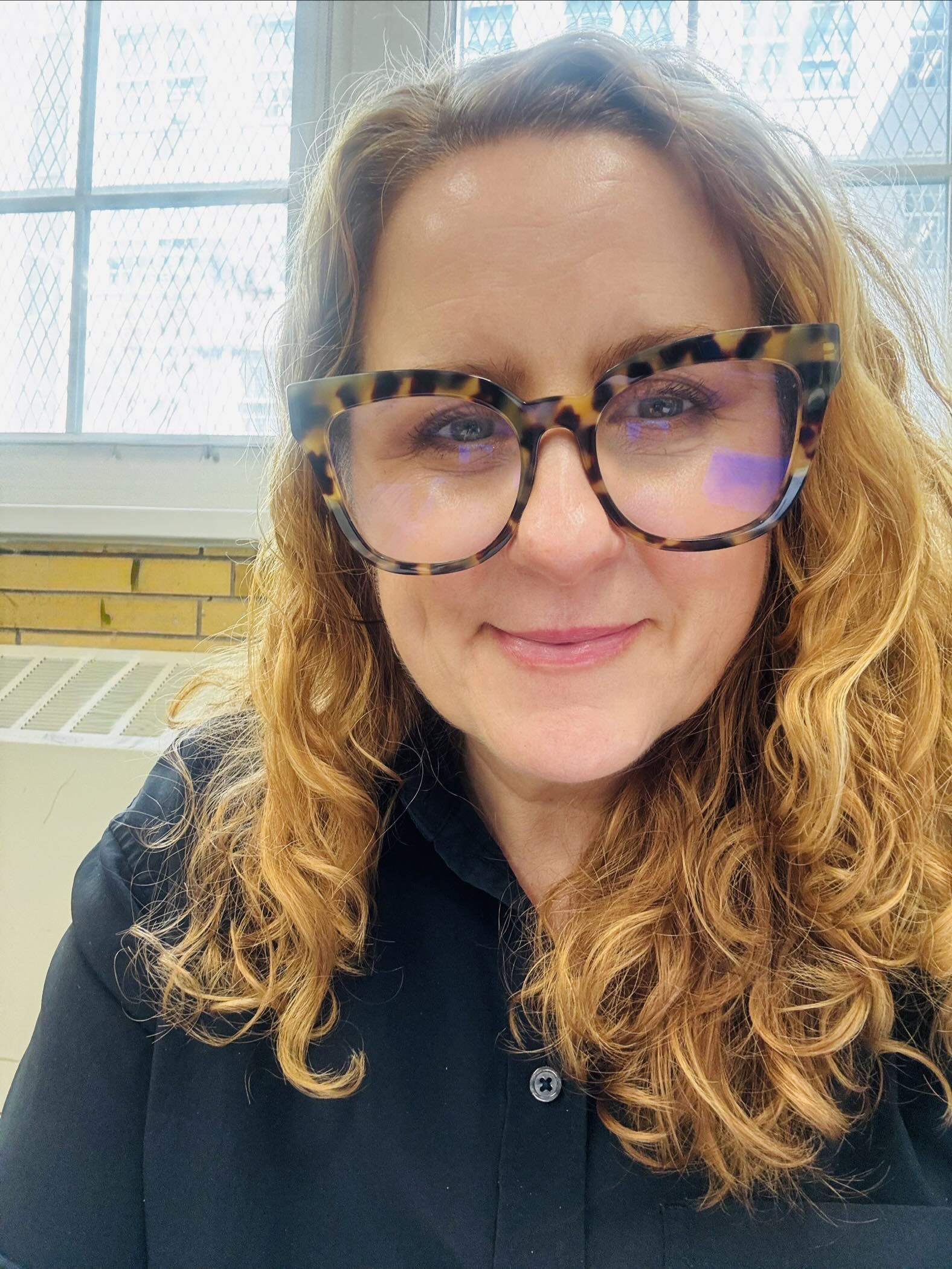 K. A. Keener, an educator with curly auburn hair and tortoiseshell glasses, in a classroom by tall wired-glass windows.